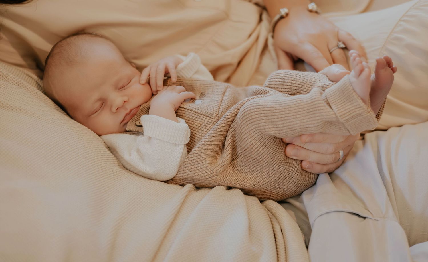 A newborn baby sleeps peacefully on cream-colored bedding while being gently cradled by caring hands.