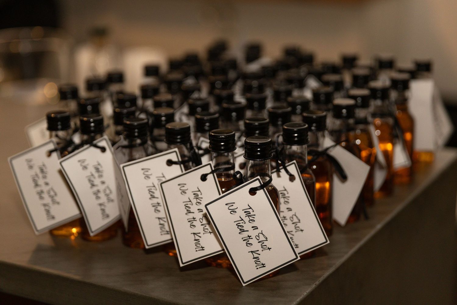 Rows of mini whiskey bottles with handwritten gift tags arranged as wedding favors on a table.