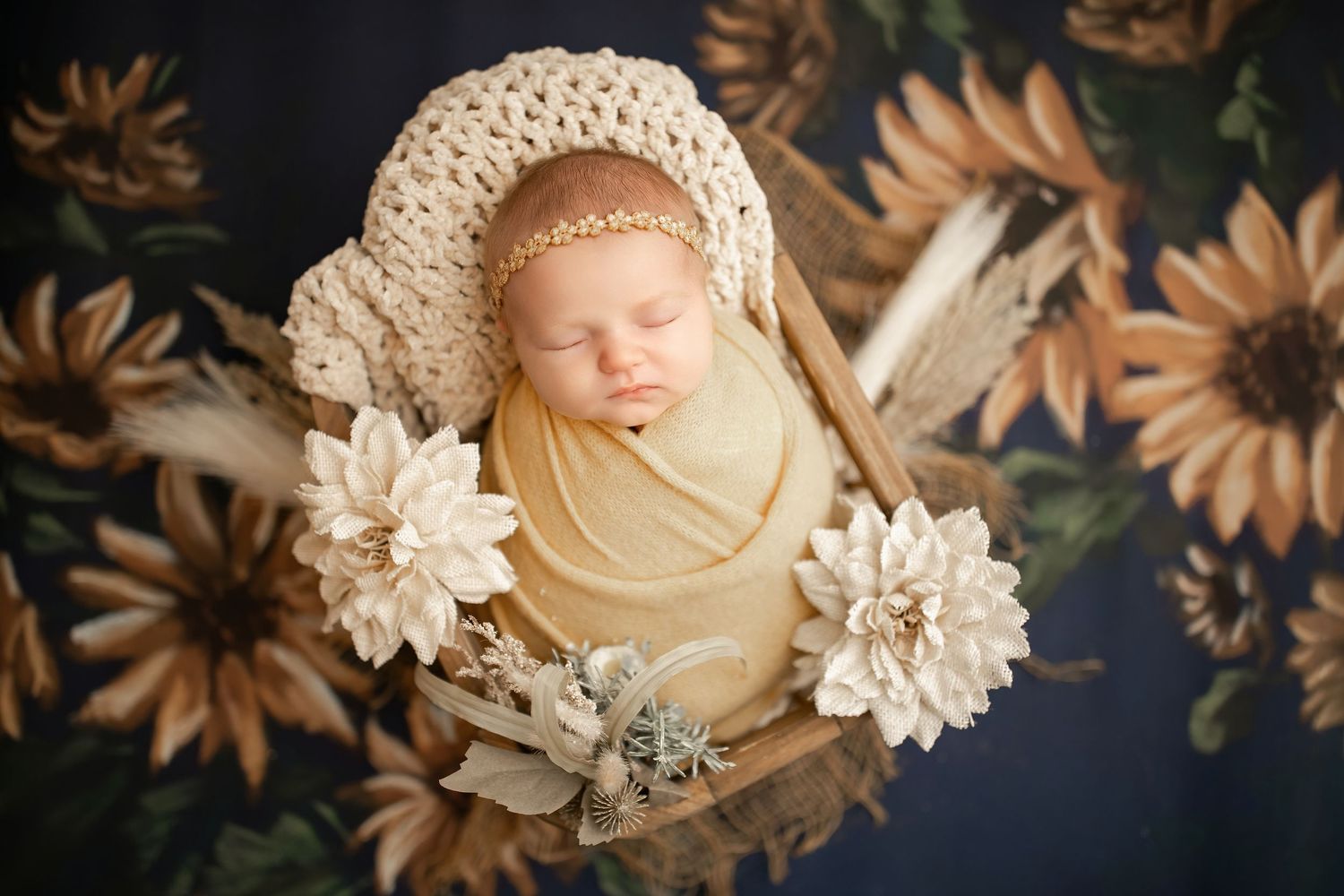 Newborn baby swaddled in yellow wrap sleeping in wooden bowl with white flowers and floral backdrop.