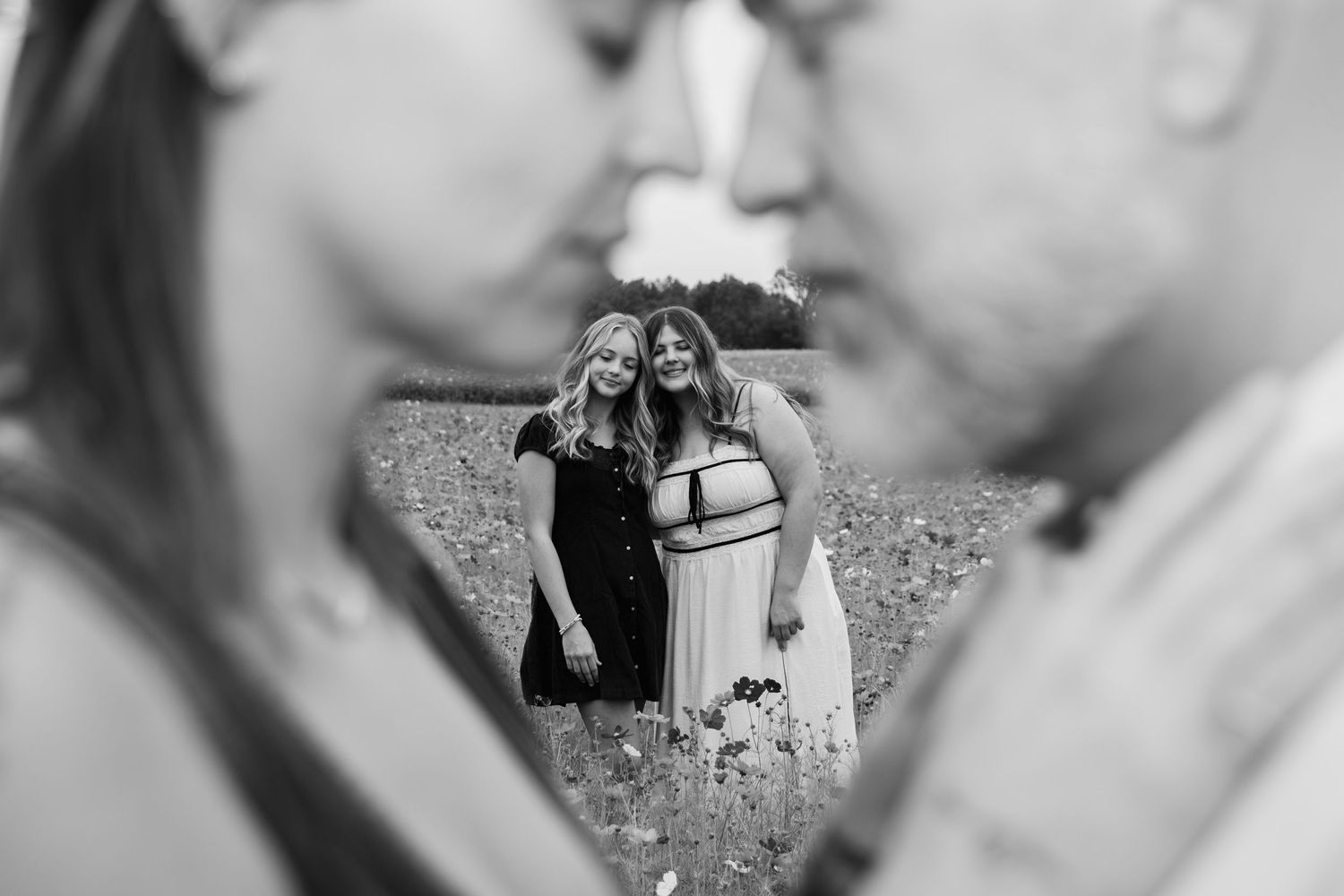 Black and white photo of two women embracing in a field, framed between the blurred faces of a couple in the foreground.