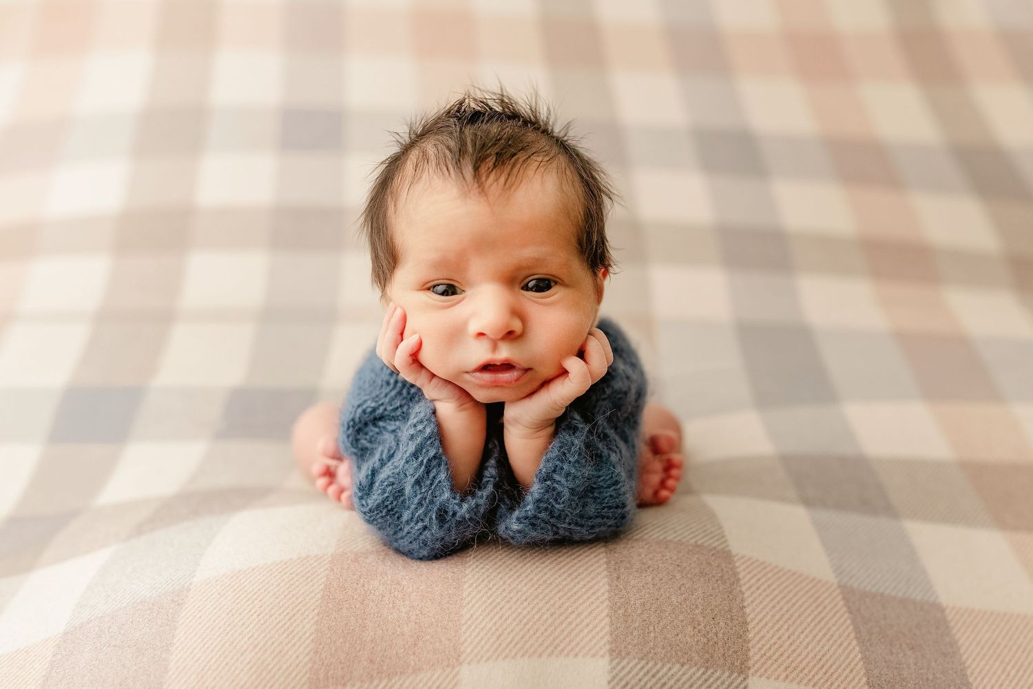 Newborn in blue knit outfit lying on plaid blanket with hands under chin.