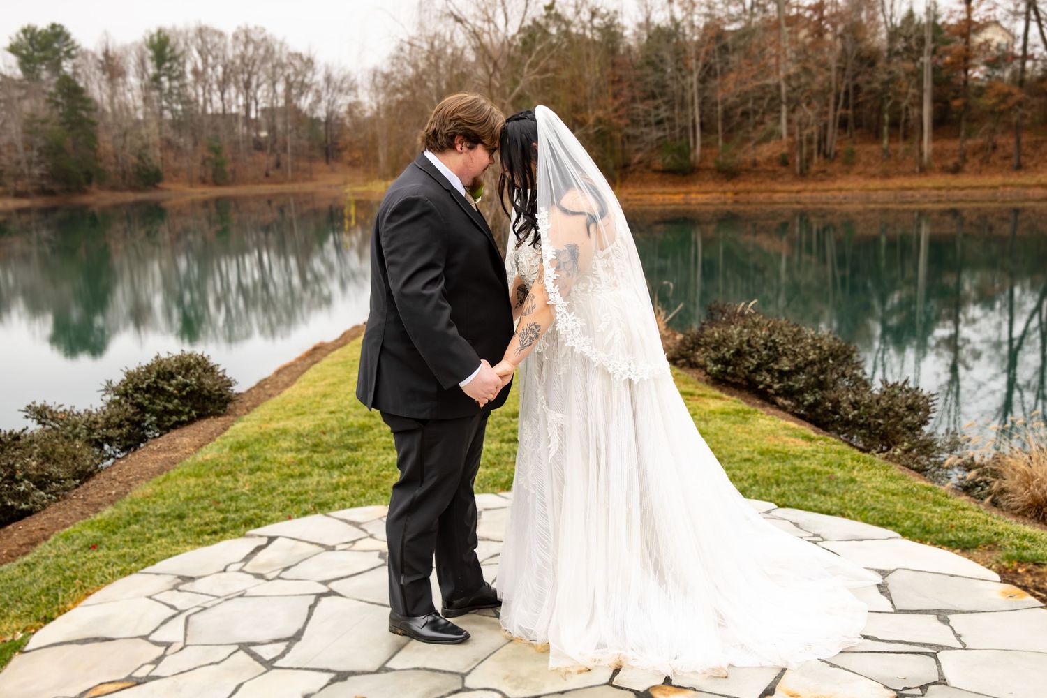Bride and groom hold hands facing each other on stone patio with tranquil lake backdrop in winter.