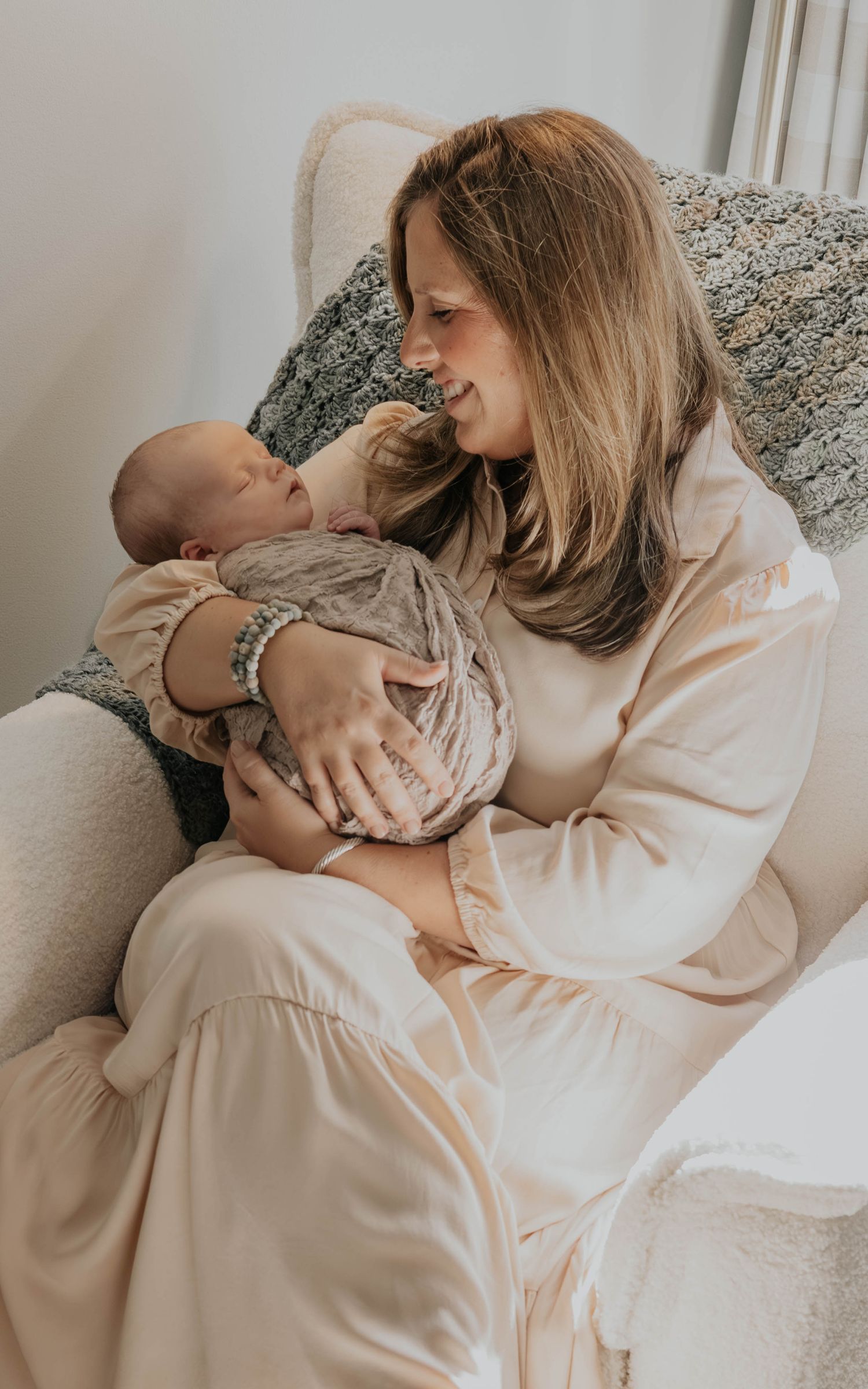 A tender moment between parent and newborn baby while sitting in a cozy armchair wearing soft neutral colors.
