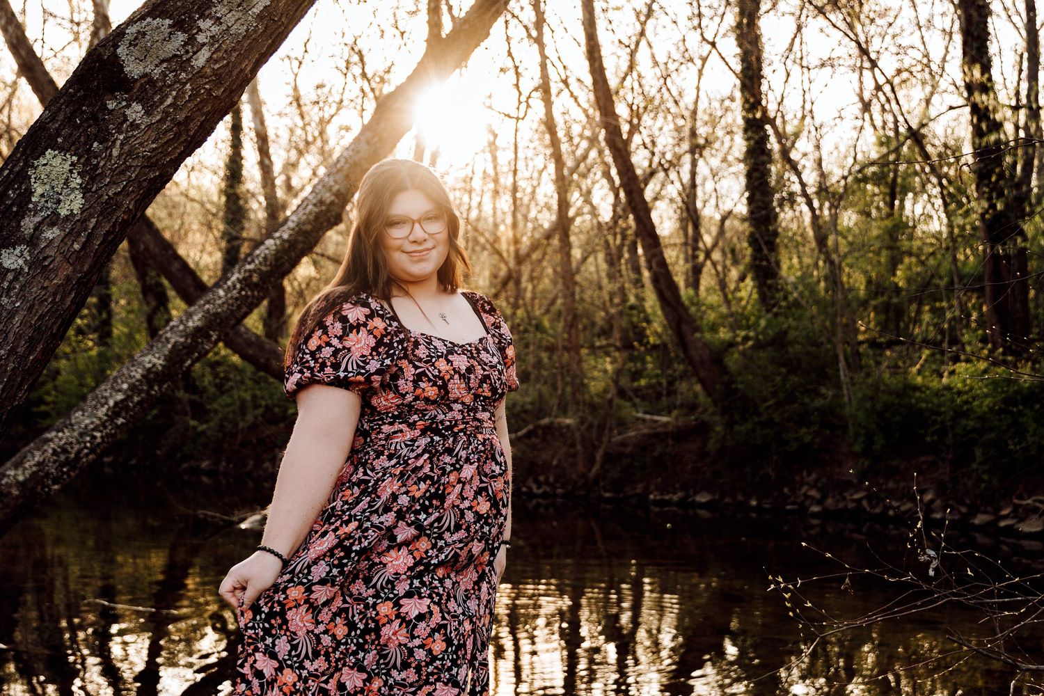 Young woman in floral dress standing by a reflective creek in a sunlit forest during golden hour.