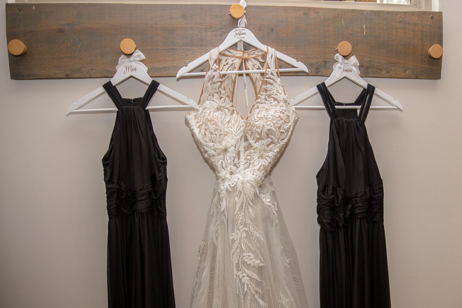 Wedding gown and two black bridesmaid dresses hanging on a rustic wooden wall rack with white hangers.