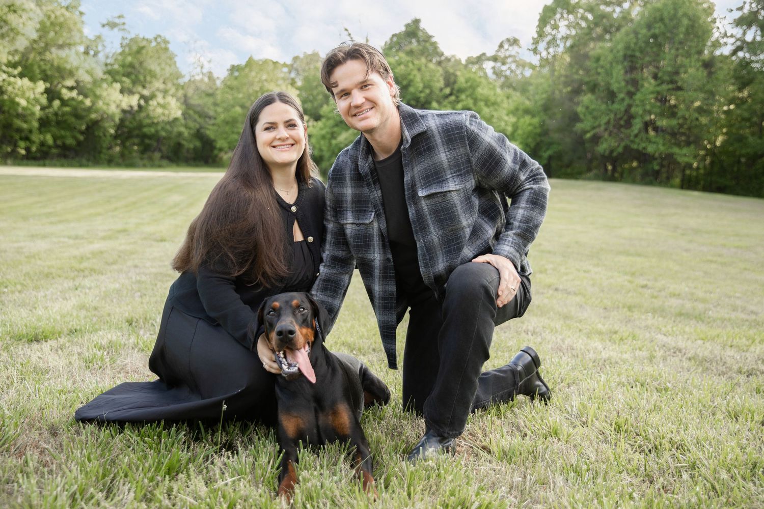Couple smiling with their Doberman dog on a green grassy field surrounded by trees.