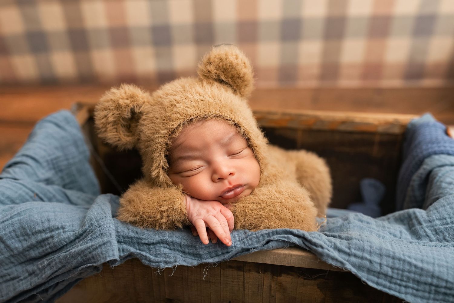 Newborn baby sleeping peacefully in a wooden crate wearing a brown teddy bear bonnet with blue fabric.