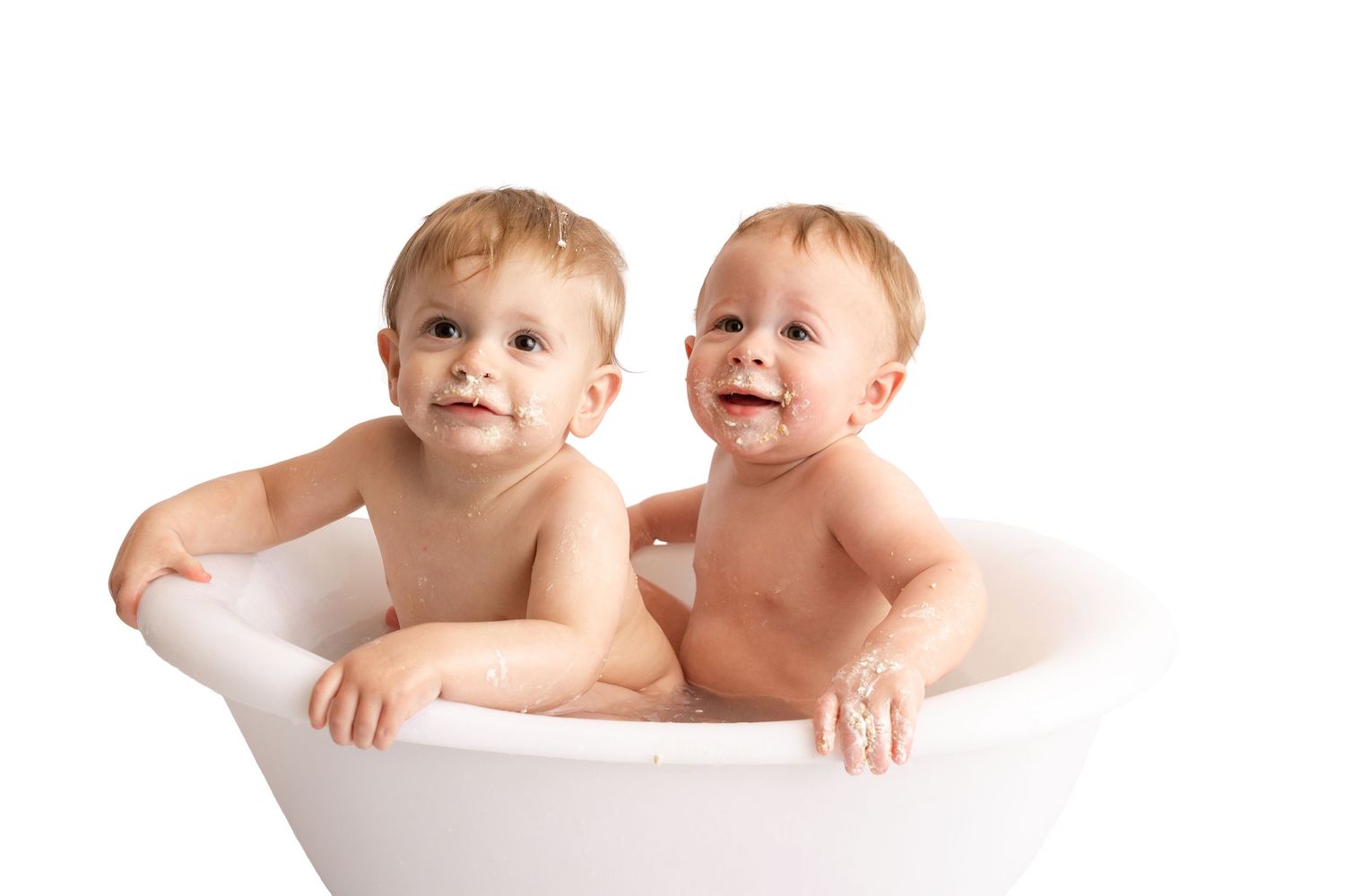 Two smiling baby boys with light brown hair sit together in a white bathtub against a clean white background.