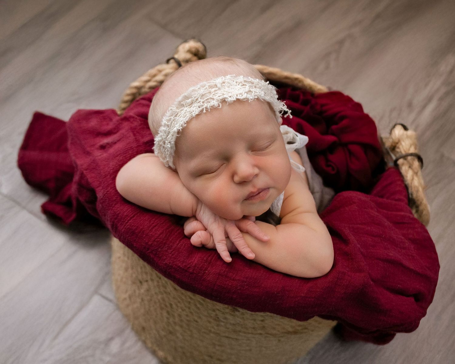 A sleeping newborn wrapped in burgundy fabric rests peacefully in a rustic basket with a delicate lace headband.