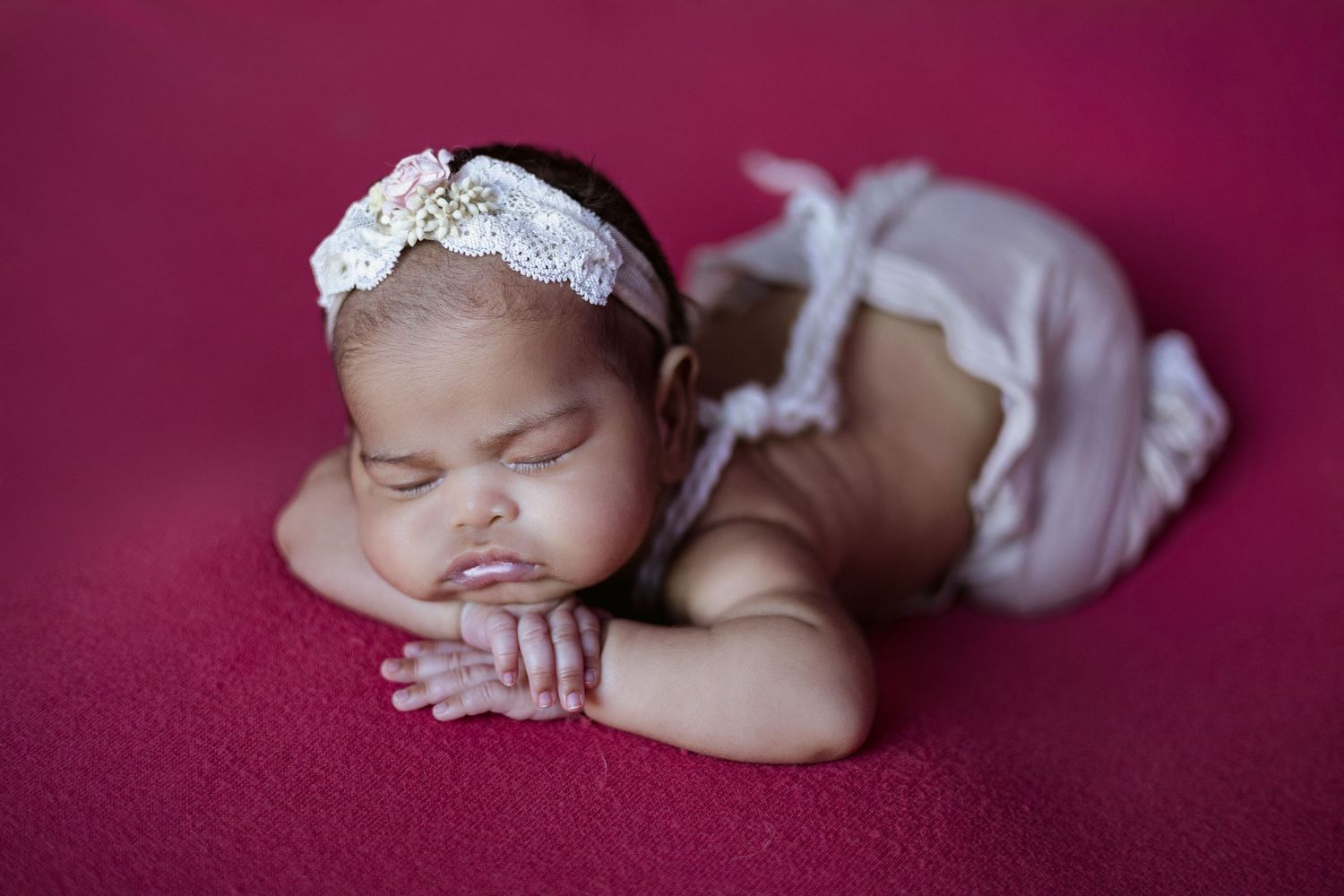 A sleeping newborn baby wearing white lace lies on a deep burgundy velvet backdrop.