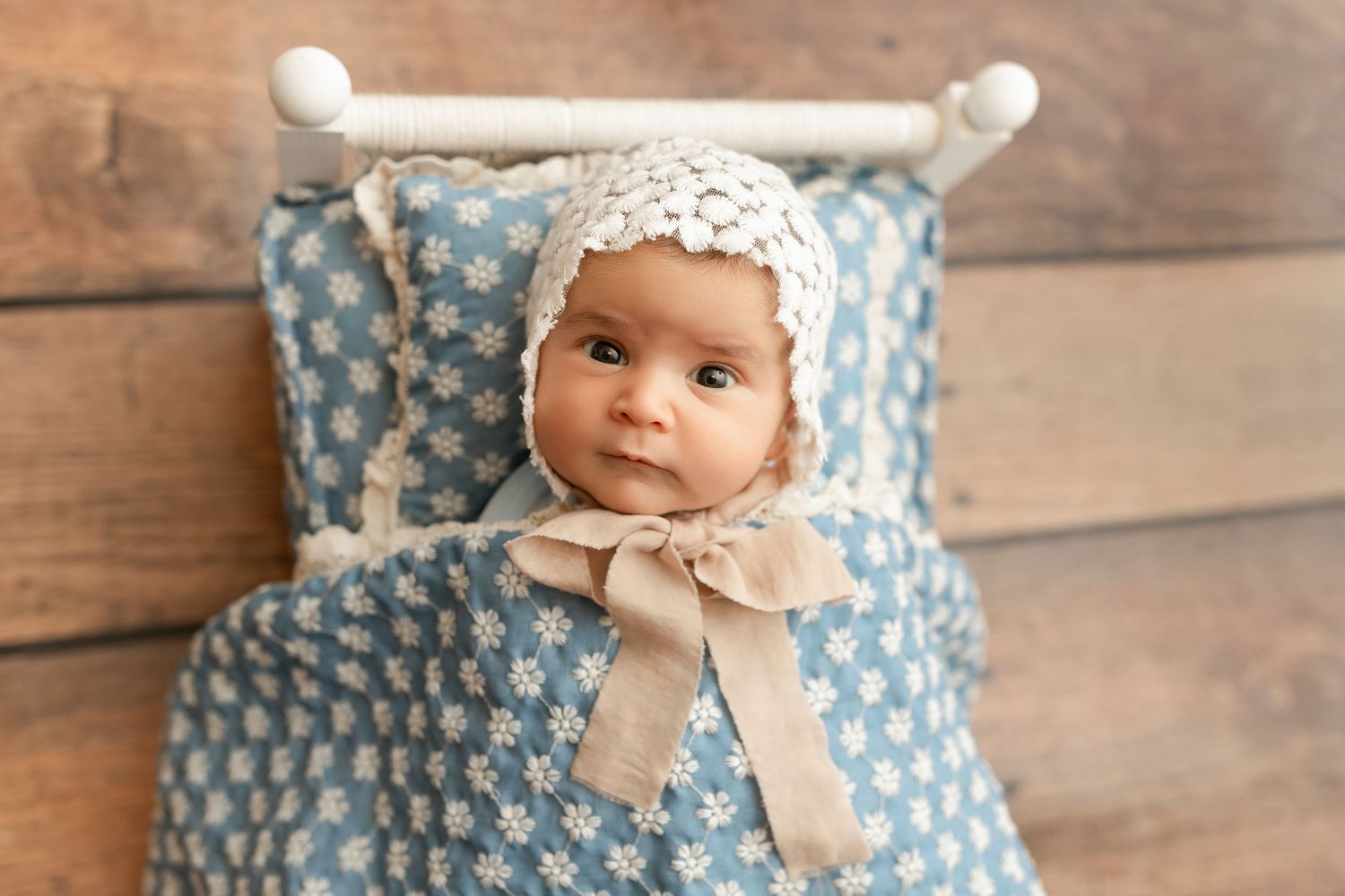 Newborn baby in a lace bonnet wrapped in a blue floral blanket lying in a small white decorative bed.