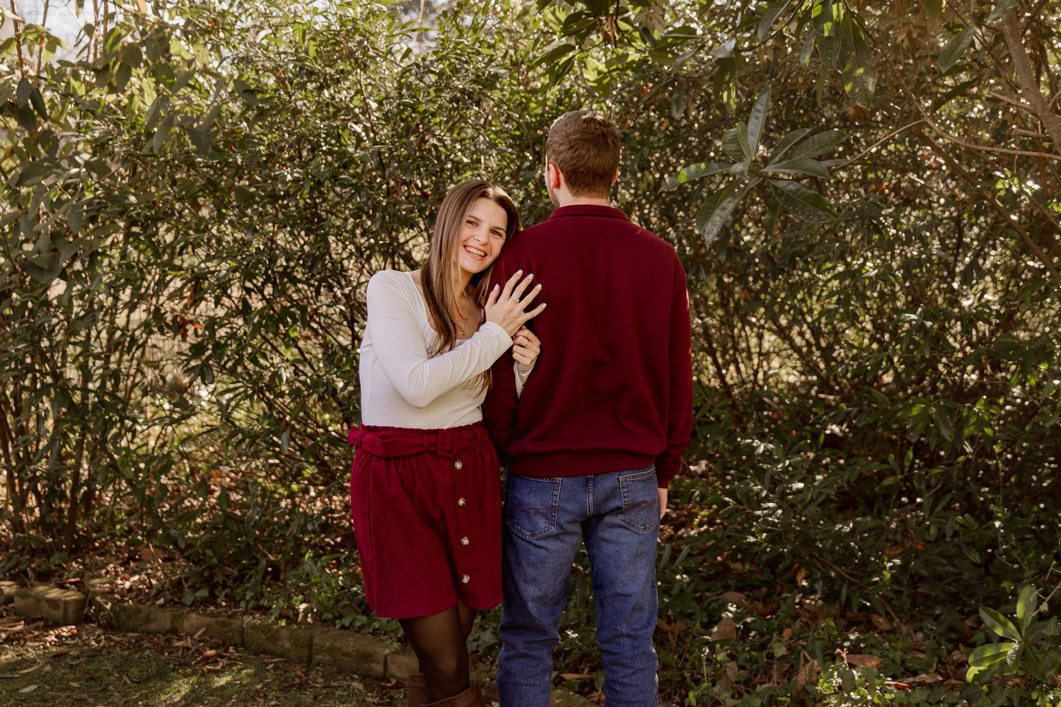 A romantic couple in burgundy and denim embraces while standing in front of lush green foliage.