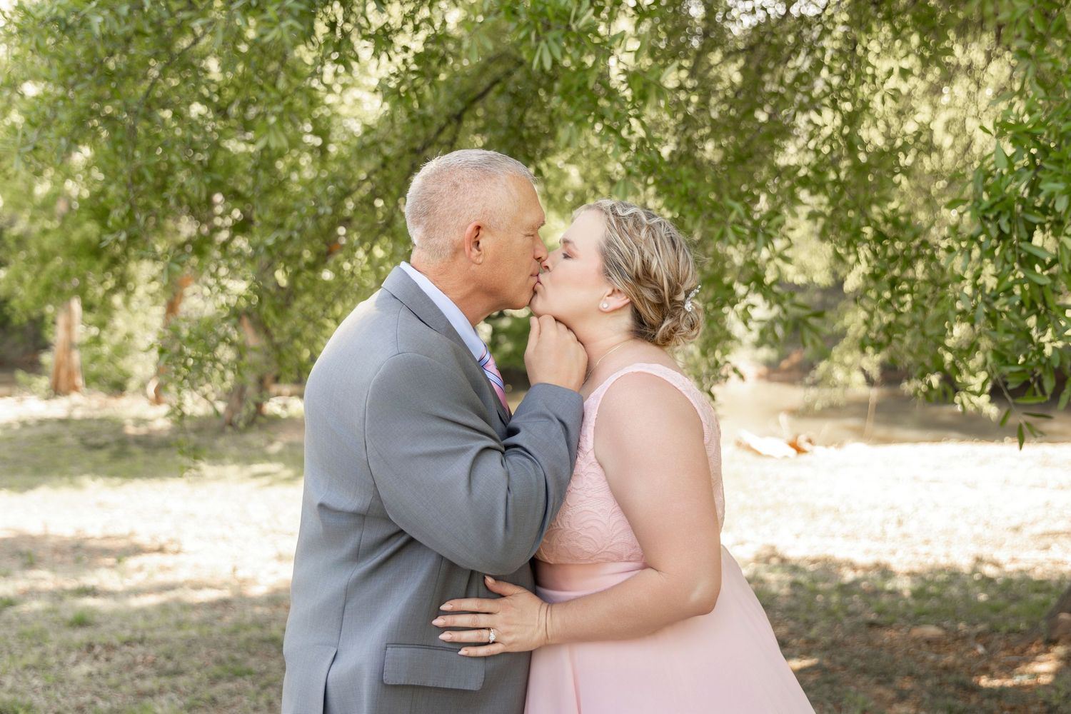 Couple sharing a romantic kiss outdoors under green trees, man in gray suit and woman in pink dress.