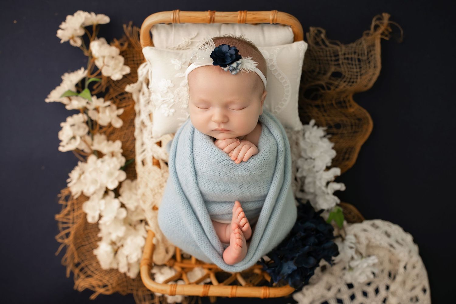 Newborn baby swaddled in blue wrap sleeping peacefully on a tiny wicker bed with white and navy floral decor.