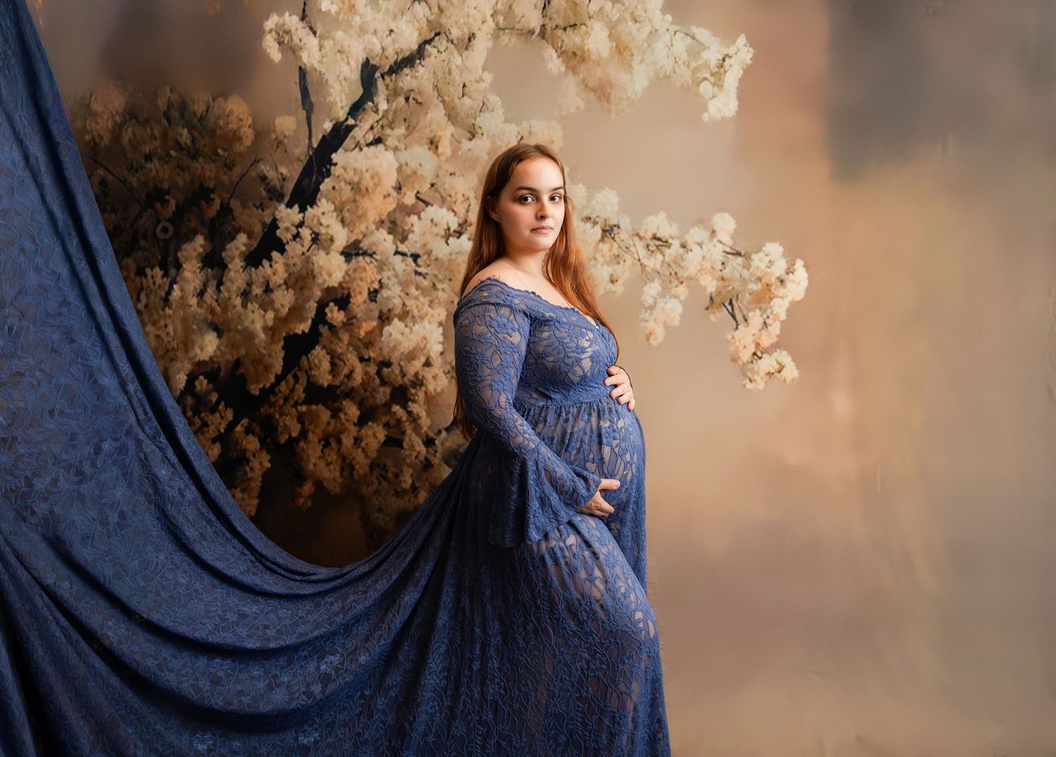 Pregnant woman in blue lace gown poses elegantly beside white floral branches in a dreamy maternity portrait.