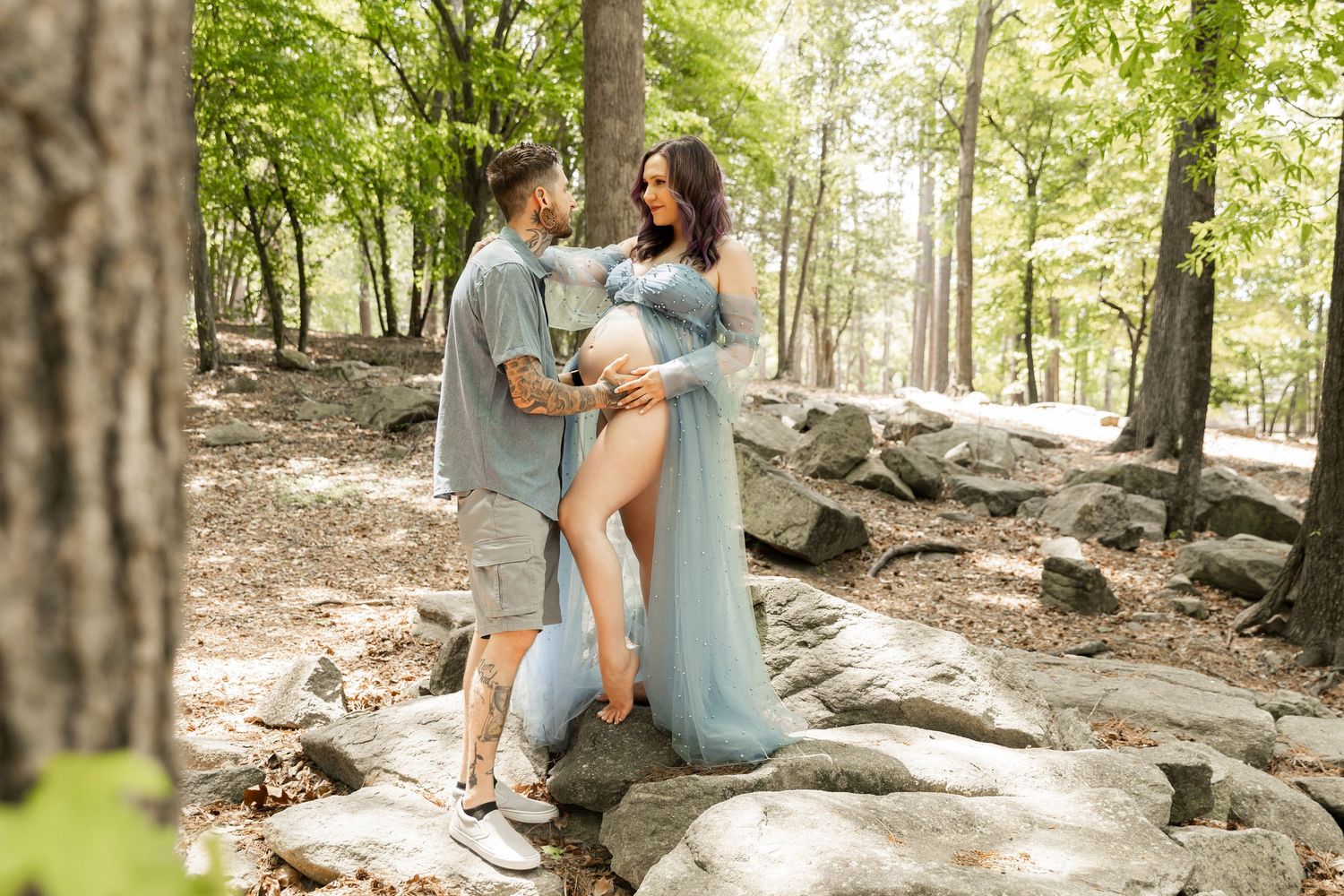Couple poses for maternity photos in a forest, woman in flowing teal gown, man in blue shirt, surrounded by rocks.