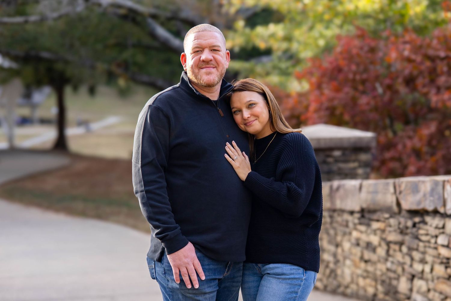 A couple poses together outdoors in autumn with colorful fall foliage and a stone wall in the background.