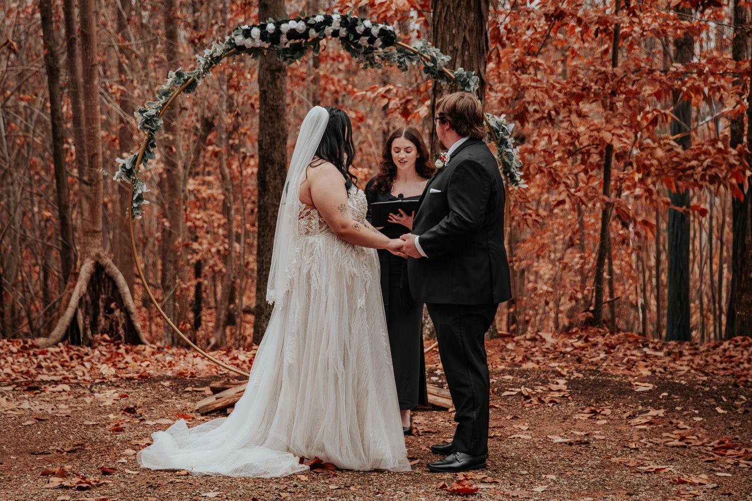 Couple exchanges vows at outdoor fall forest wedding ceremony beneath floral arch with officiant.