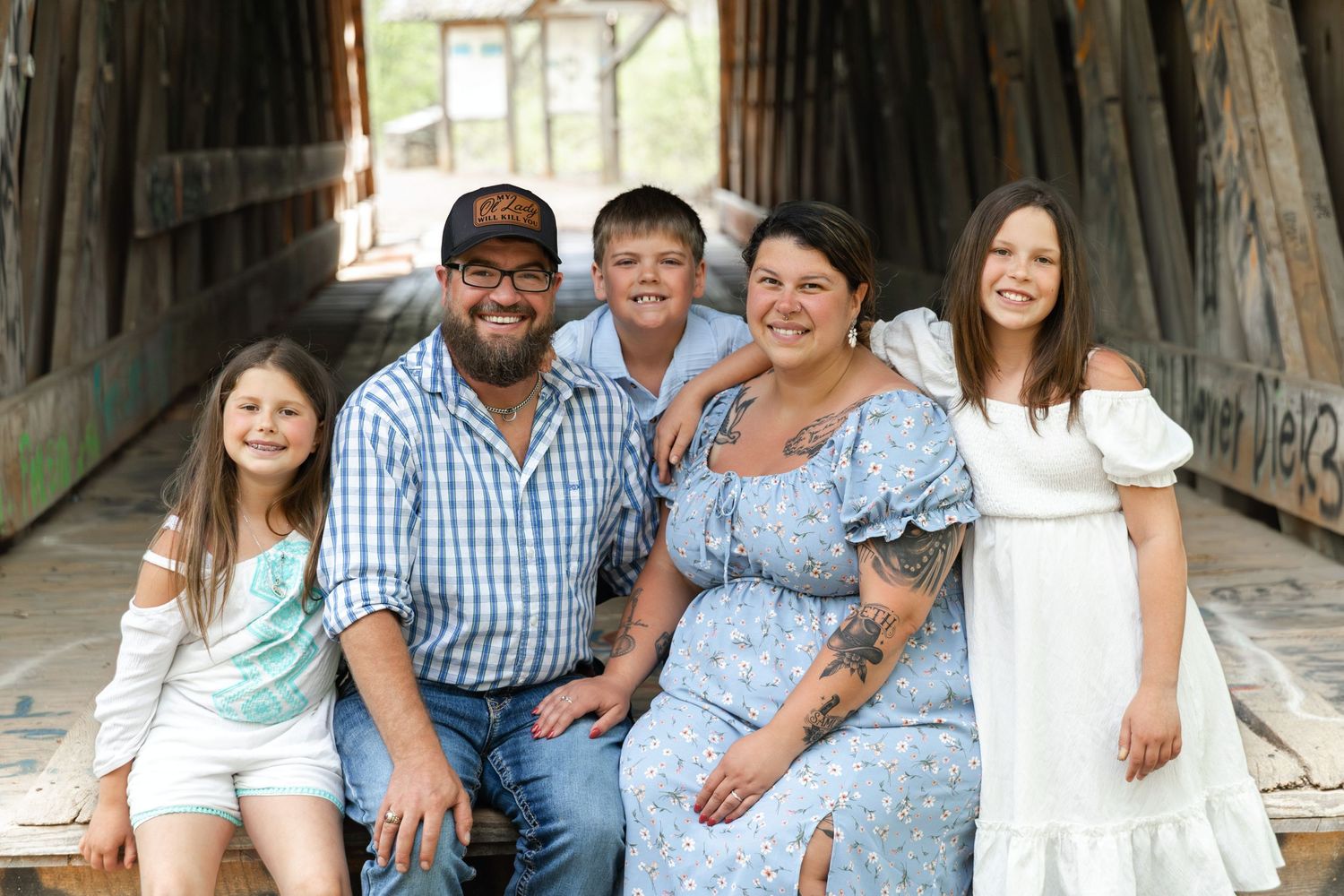 A happy family of five smiling together inside a rustic covered bridge, dressed in casual summer outfits.