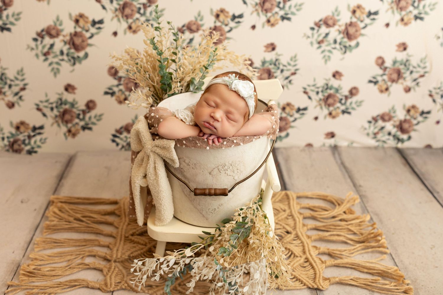 Newborn baby girl sleeping peacefully in a cream bucket with floral decor and a delicate bonnet on a wooden floor.