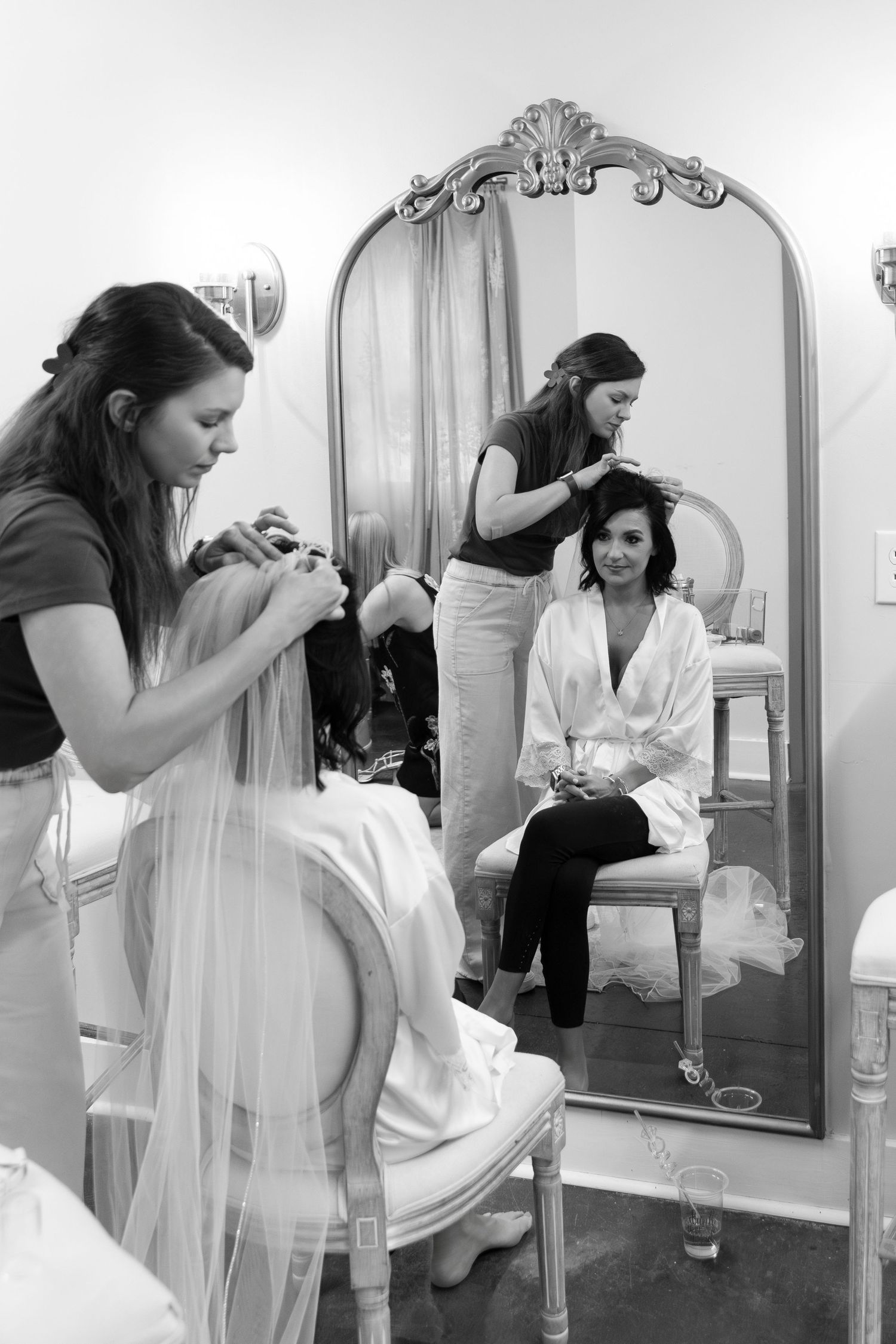 Bride getting hair styled by two assistants in a elegant dressing room, reflected in ornate mirror, black and white photo.
