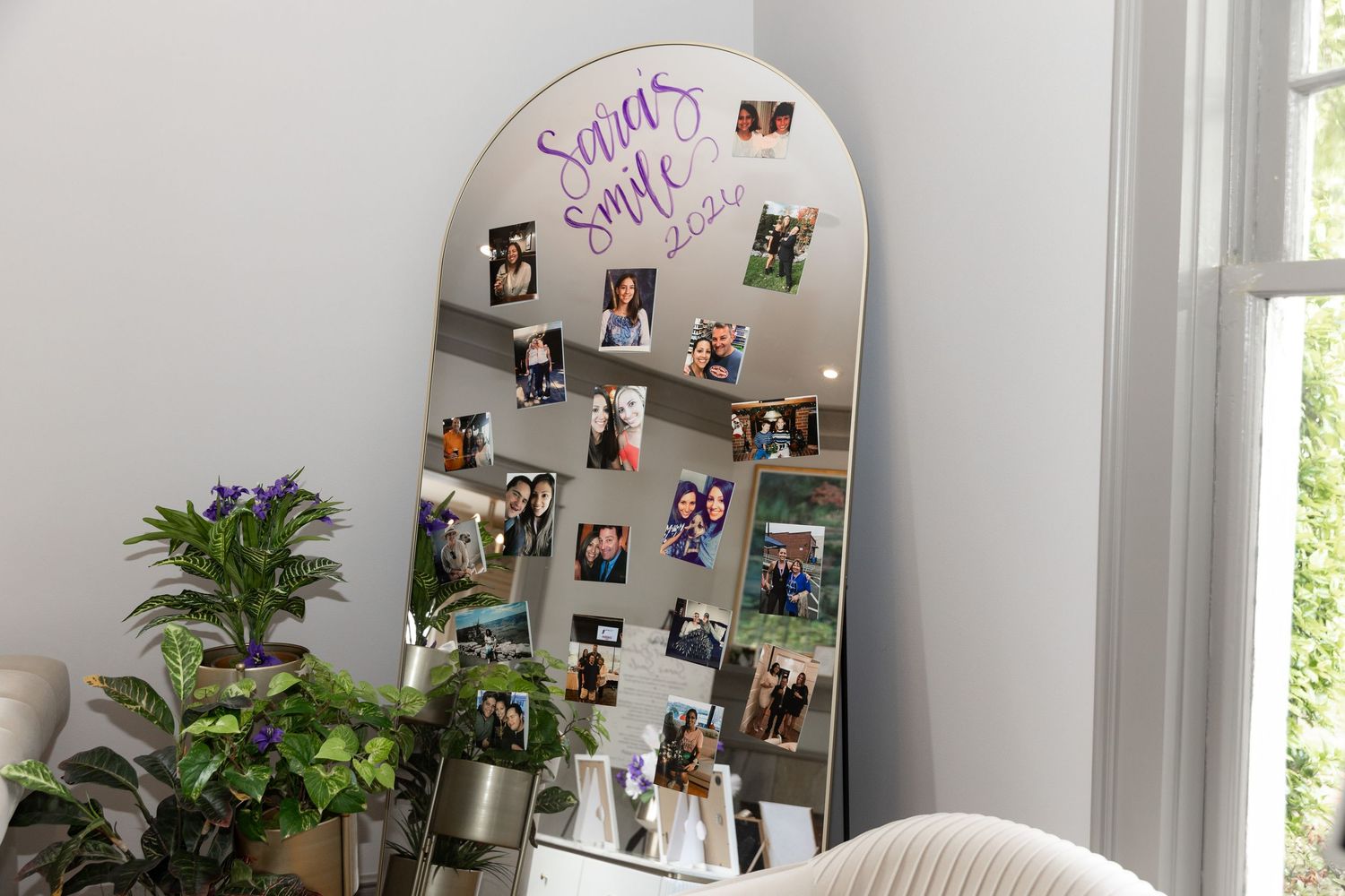 A large arched mirror leaning against a wall, decorated with photos and cursive writing, surrounded by green plants.