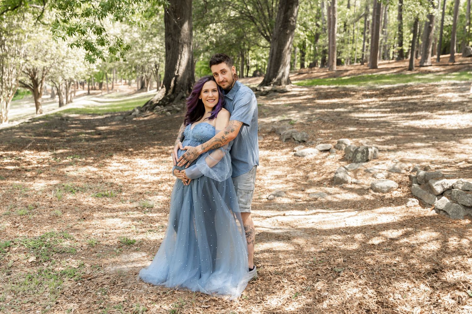 Couple posing in a forest park, woman in blue tulle gown held by man in casual attire, maternity photo session.
