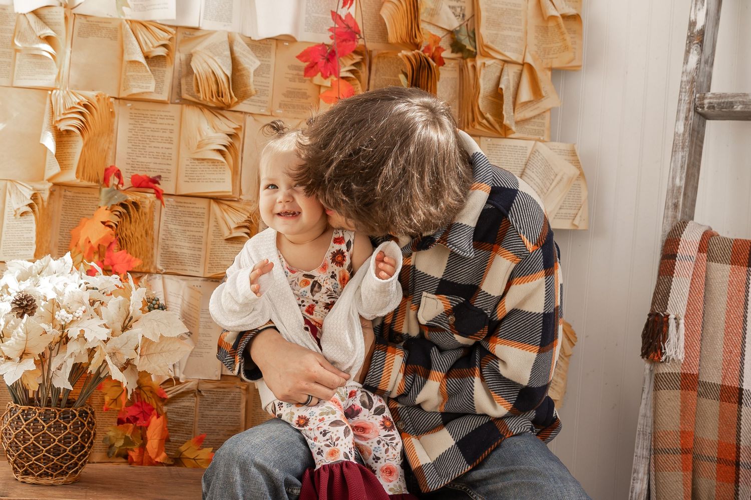 Parent in a plaid flannel shirt cuddles a smiling toddler in a cozy autumn family photo setting with fall leaves and a rustic wall backdrop.