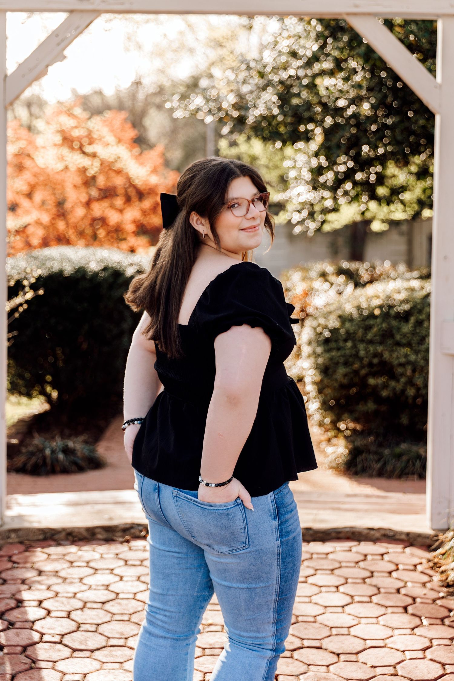 Young woman in black top and jeans poses in a sunlit garden with lush greenery and brick pathway.