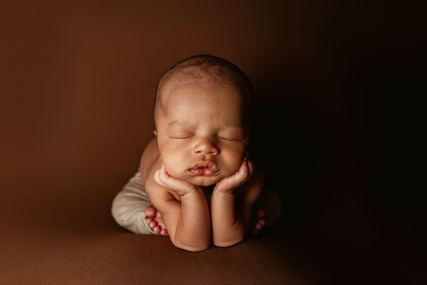 A sleeping baby rests their head on folded hands against a brown background in a peaceful newborn portrait.