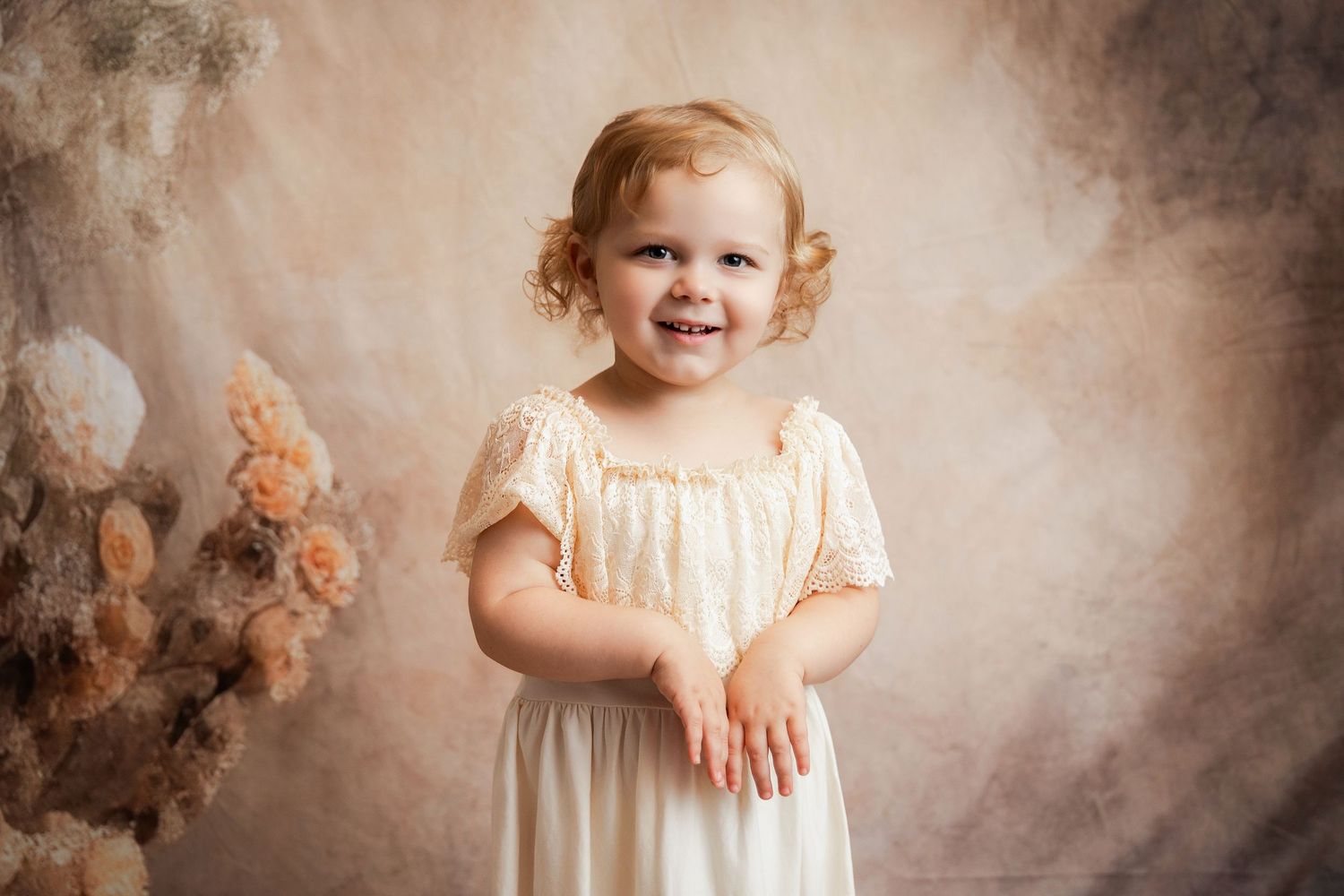 Smiling toddler girl in cream dress poses against soft floral backdrop in professional portrait photo.