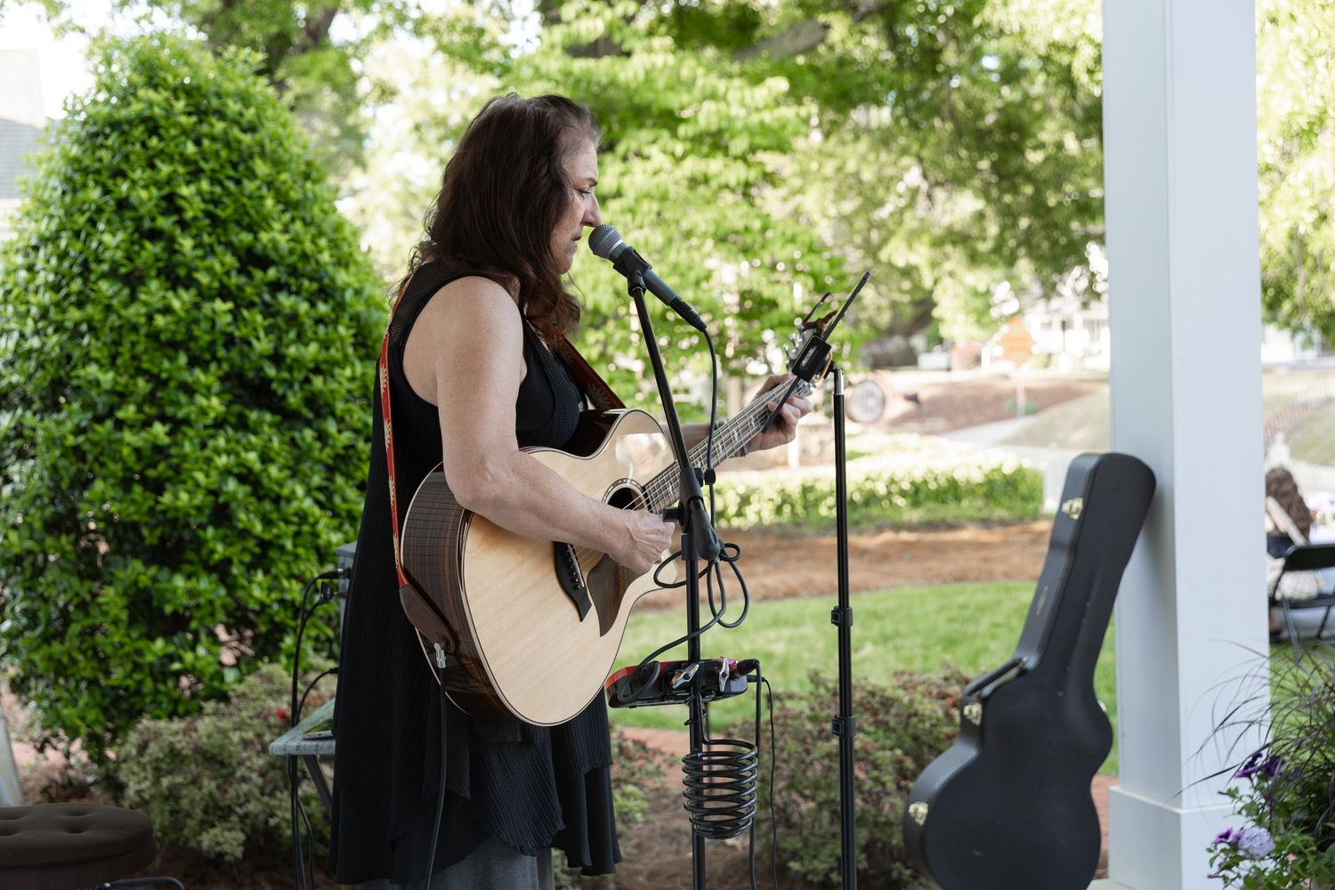Woman playing acoustic guitar and singing into microphone outdoors on porch surrounded by lush green trees.