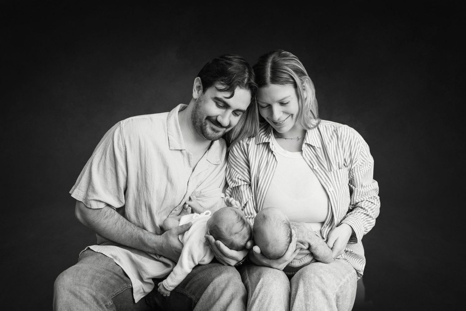 Black and white photo of smiling parents holding their newborn twins in a studio portrait.