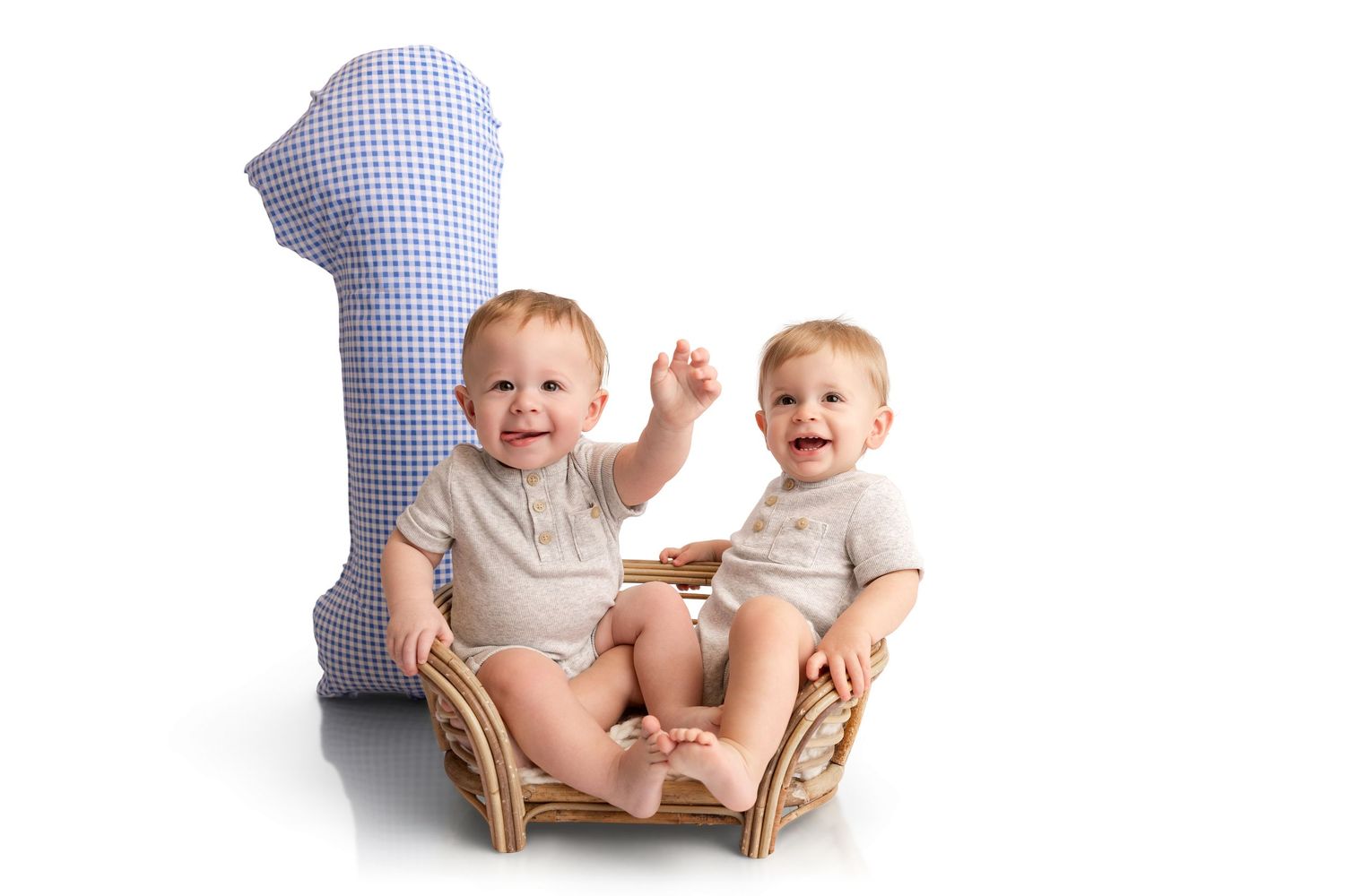 Two smiling baby boys in matching beige outfits sit together in a small wicker chair next to a large blue gingham number one pillow.