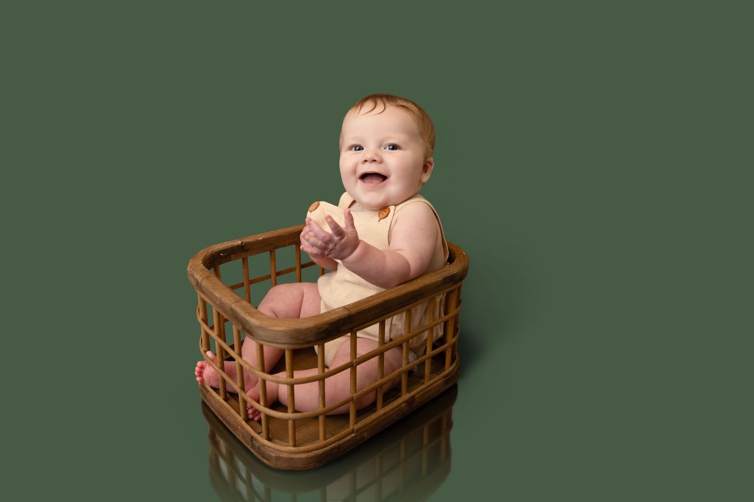 Happy baby sitting in a wooden basket, smiling and clapping hands against a green background.