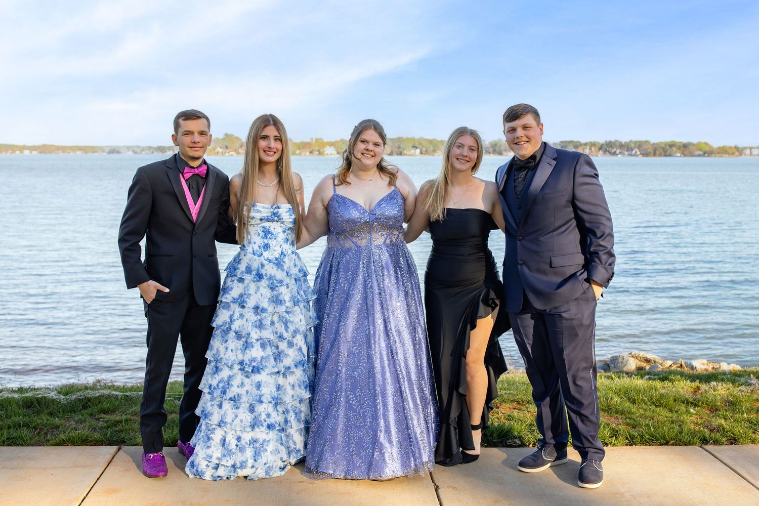 Five teens in formal prom attire pose together outdoors near a waterfront on a sunny day.