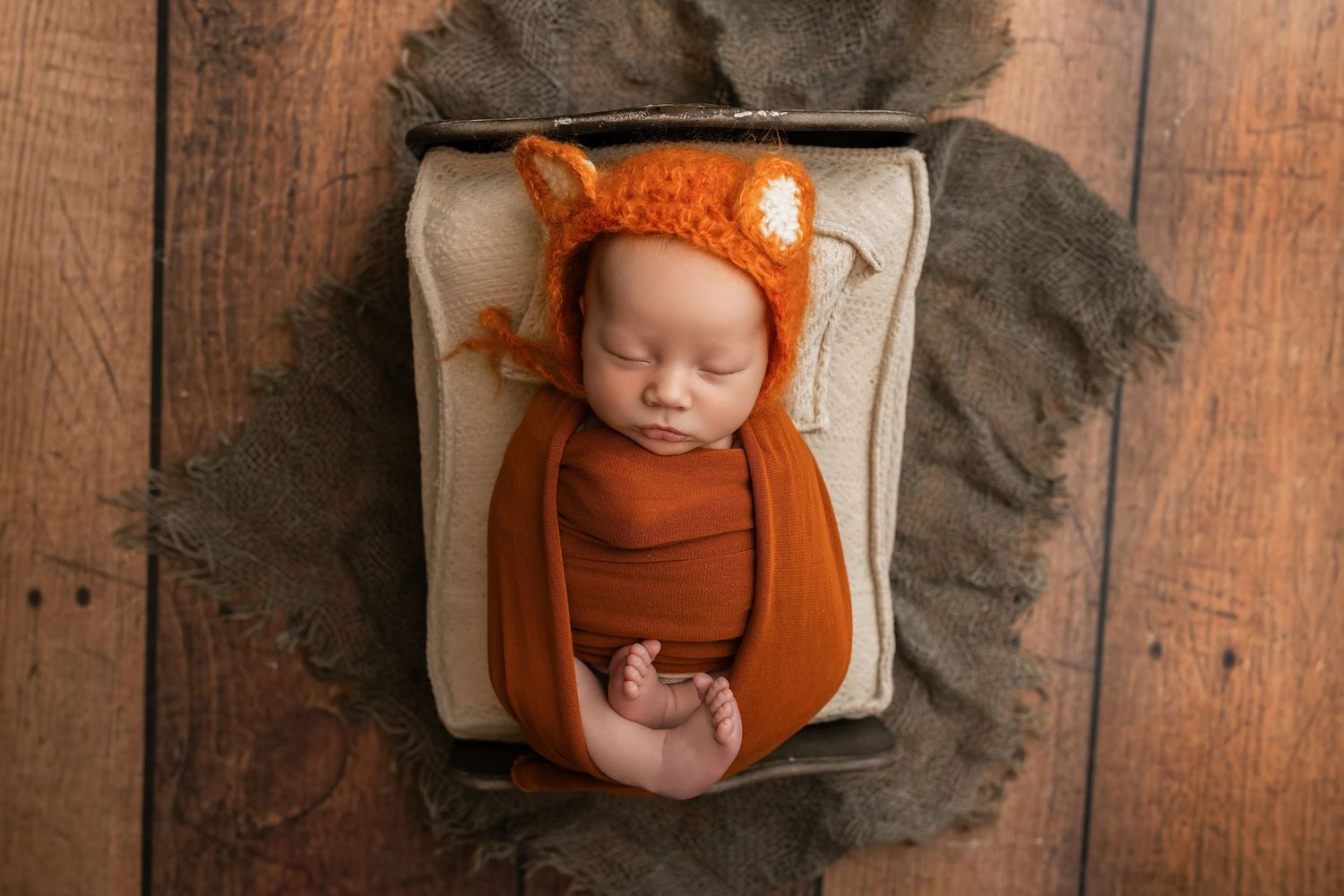Newborn baby sleeping in a wooden box wrapped in orange, wearing a knit fox bonnet hat.