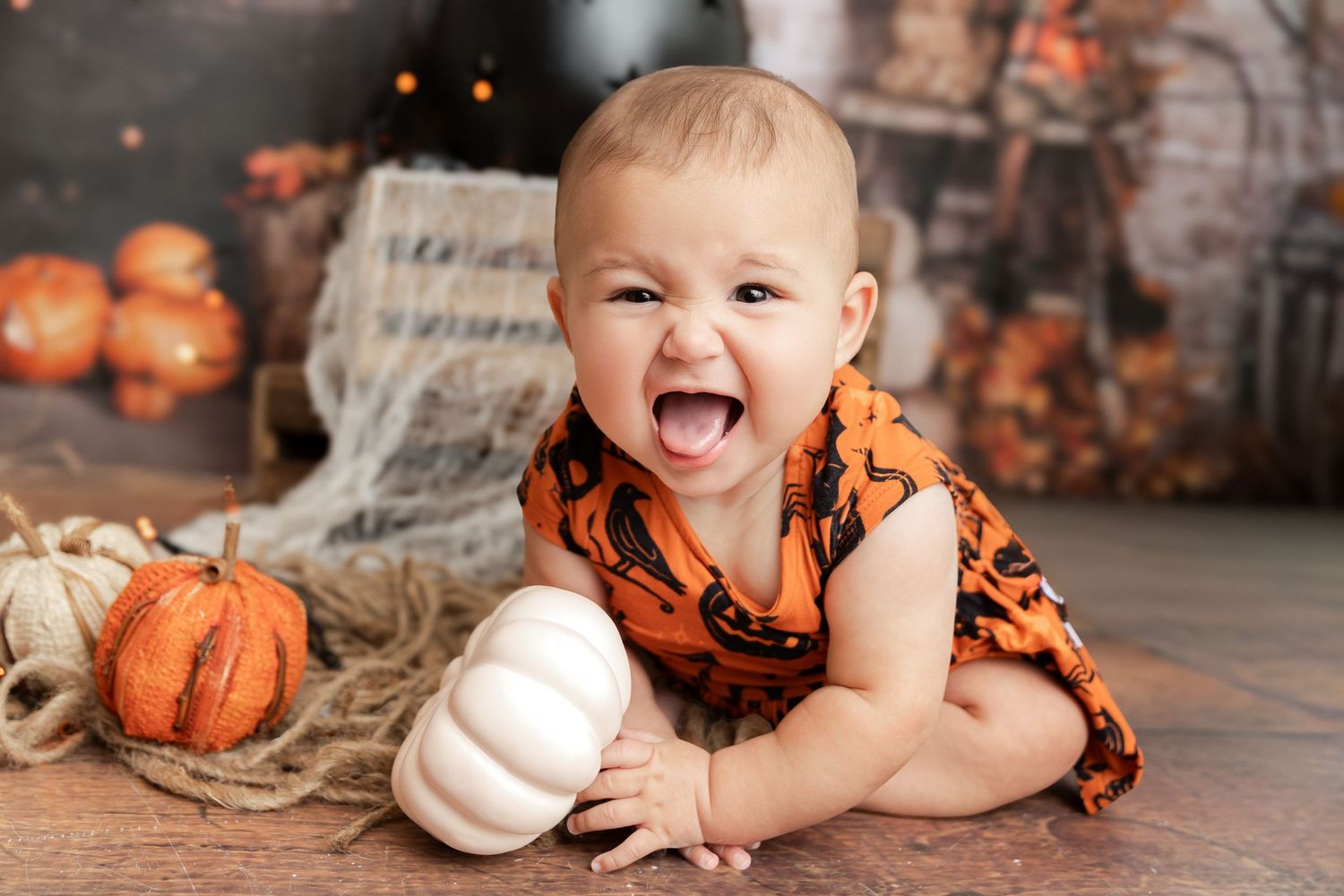 Excited baby in an orange and black Halloween outfit sits on a wooden floor beside small pumpkins and rustic burlap decor.