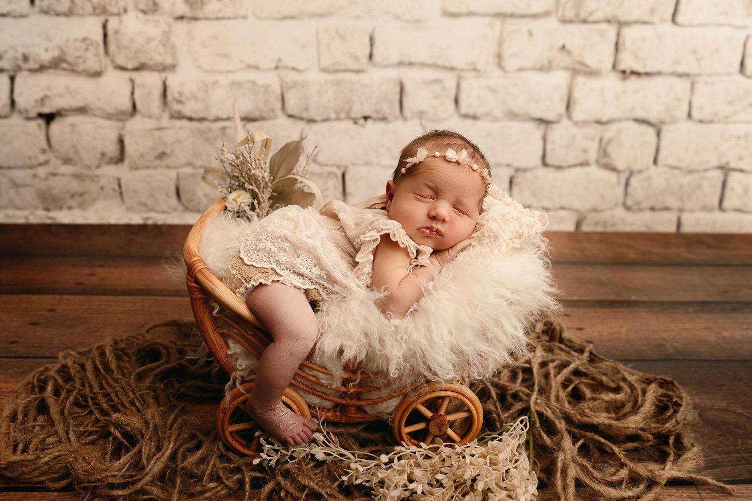 Sleeping newborn baby girl in a floral headband and lace outfit resting on a fluffy blanket in a wicker pram against a white brick wall.