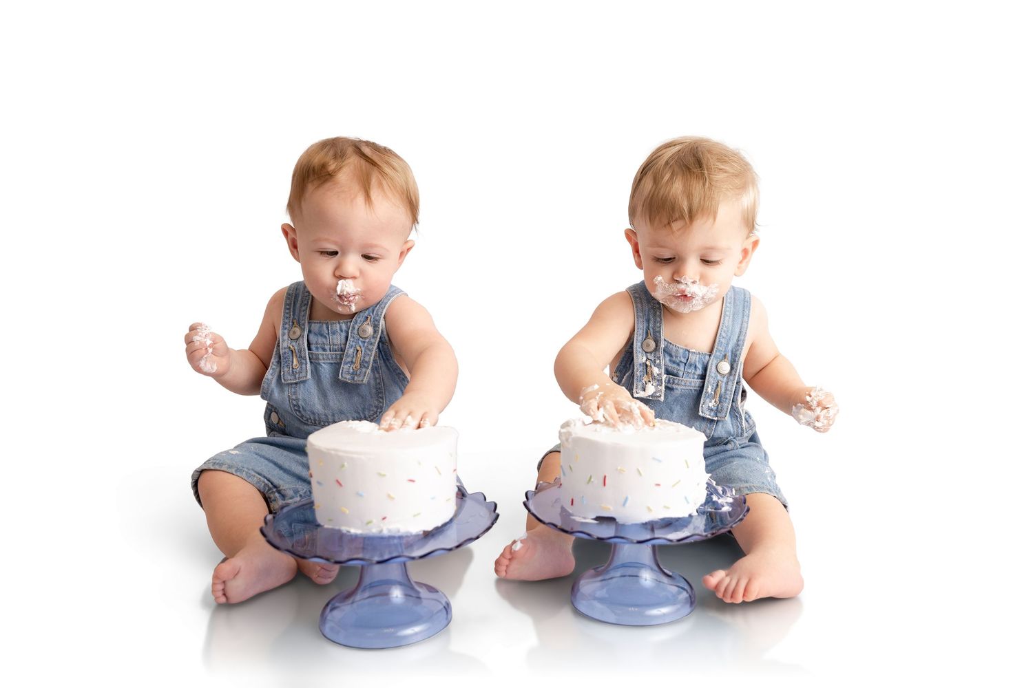 Two toddlers in denim overalls sit barefoot on a white background, each with frosting on their faces, enjoying a smash cake on a stand.