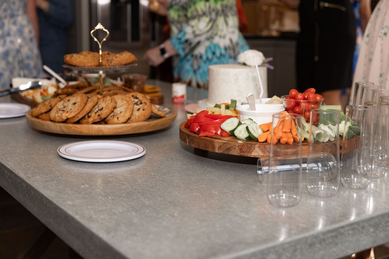 A party spread with cookies, fresh vegetables, and glasses on a gray stone countertop.