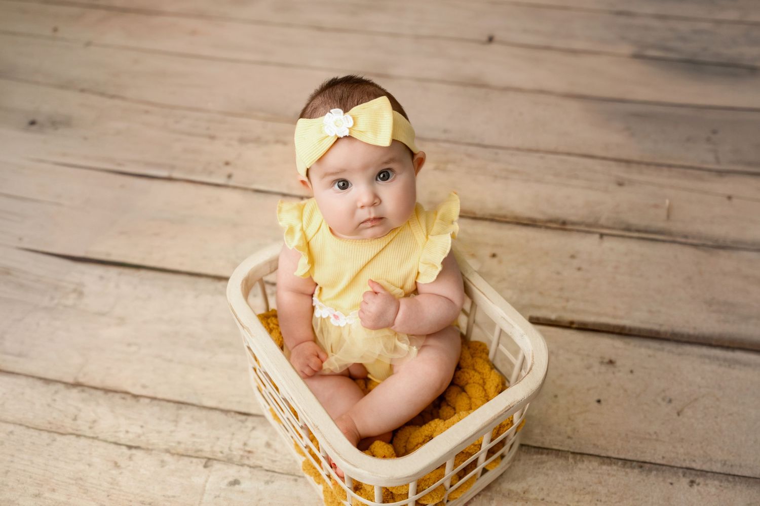 Adorable baby girl in yellow romper and bow headband sitting in a white basket on wooden floor.