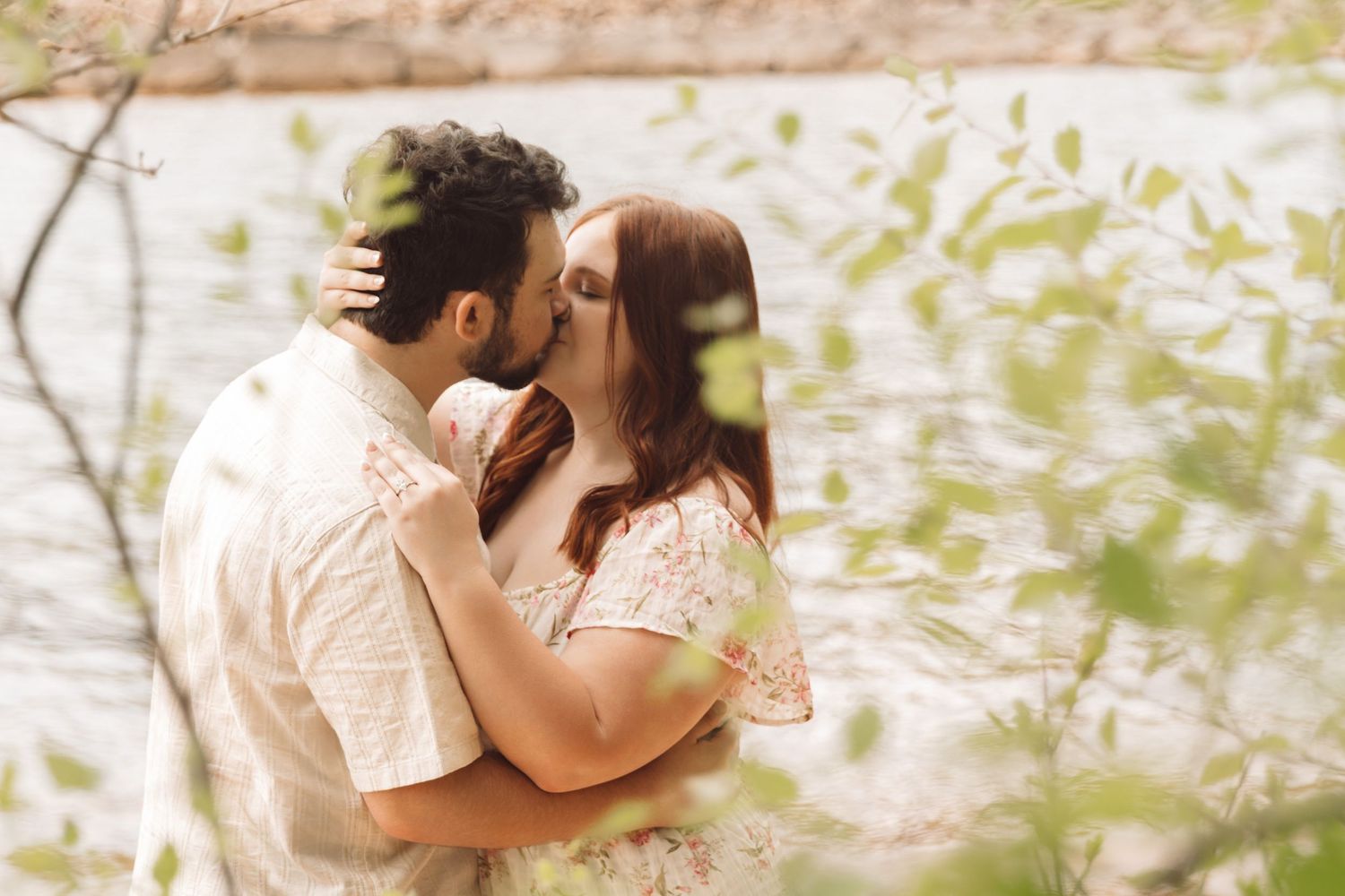 Couple sharing a romantic kiss outdoors near a lake, framed by lush green leaves in a dreamy nature setting.