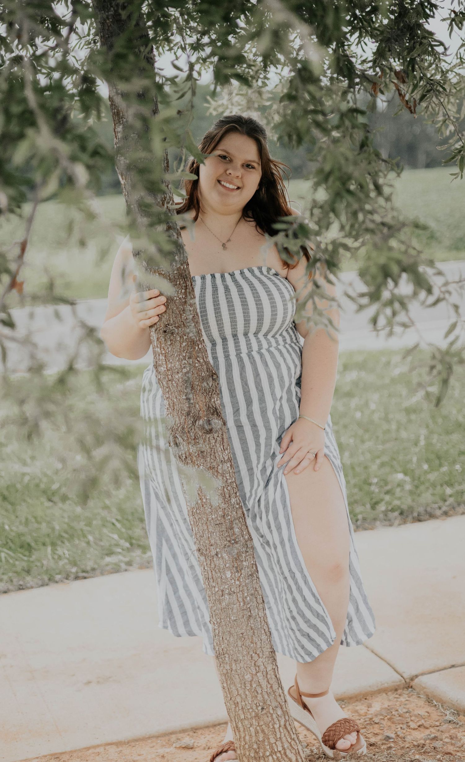 A person in a striped dress poses outdoors near a tree trunk in a dreamy, light-filled setting.