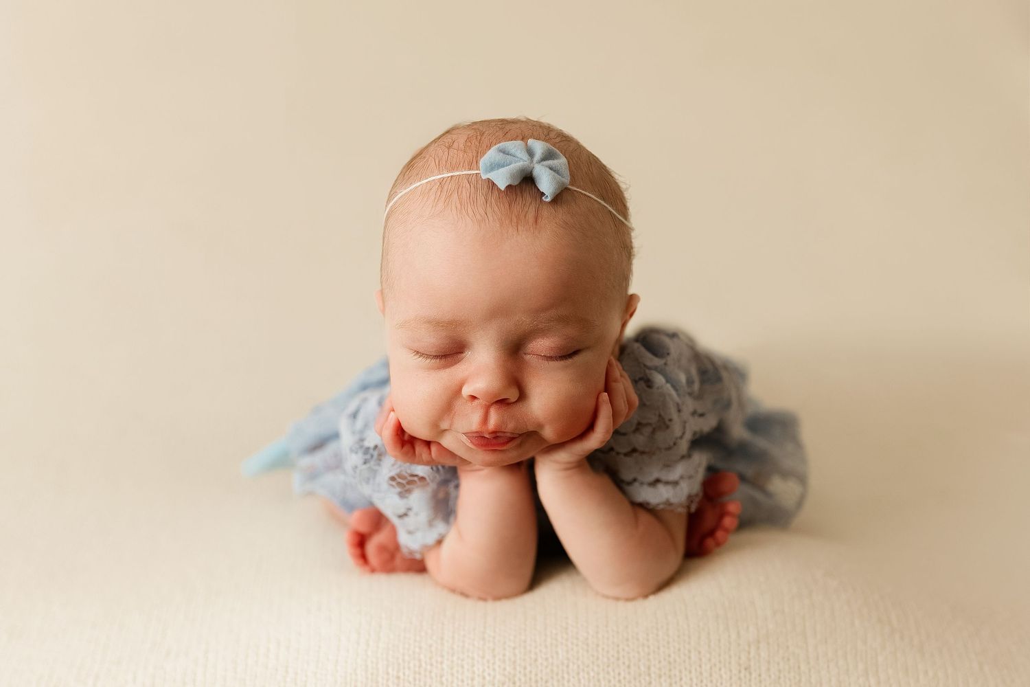A sleeping newborn baby wearing a light blue outfit rests peacefully on a cream-colored backdrop.