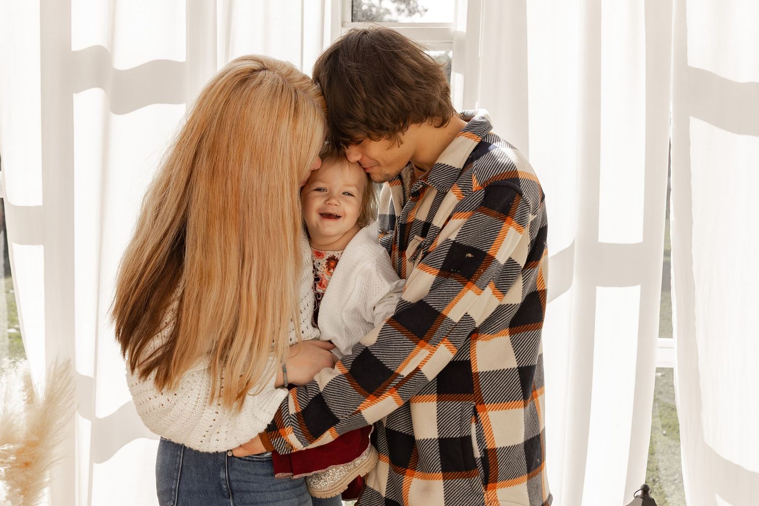Smiling family of three embracing in a bright room with white curtains, a child in the middle of a mother and father in casual plaid.