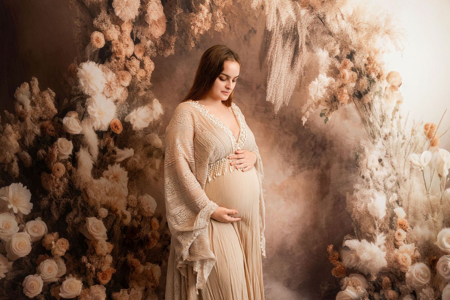 Pregnant woman in elegant beige gown poses gracefully among soft floral backdrop in a professional maternity photo shoot.