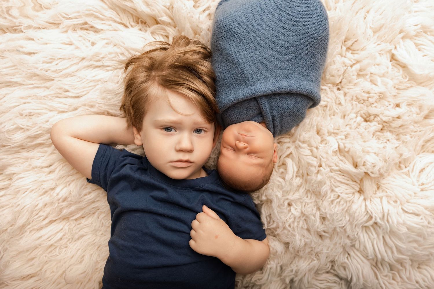 A young toddler boy lies on a fluffy cream rug beside a sleeping newborn baby wrapped in a blue swaddle.