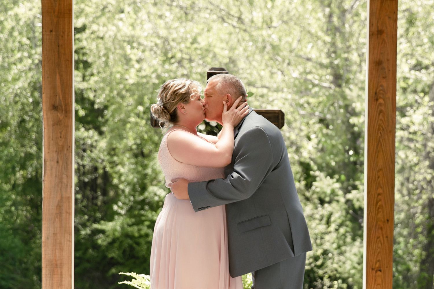Bride and groom share their first kiss at outdoor wedding ceremony surrounded by lush green trees.