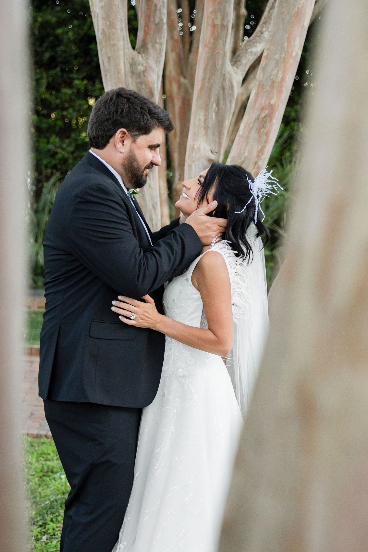 A bride and groom share a tender moment outdoors, with the groom gently cupping the bride's face near a tree.